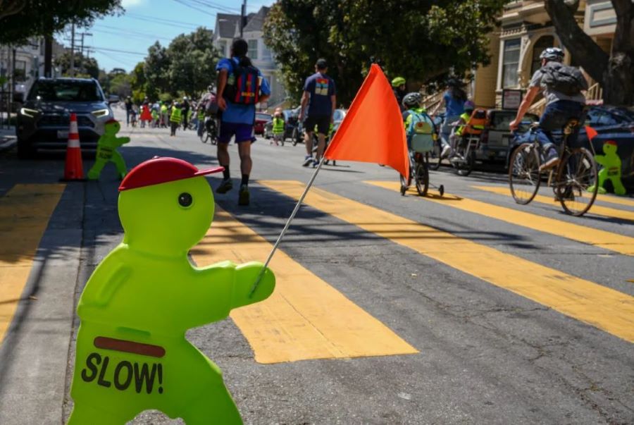 Children and adults bicycling on a street with a yellow crosswalk and a bright green plastic sign of a person that says slow and is holding an orange flag
