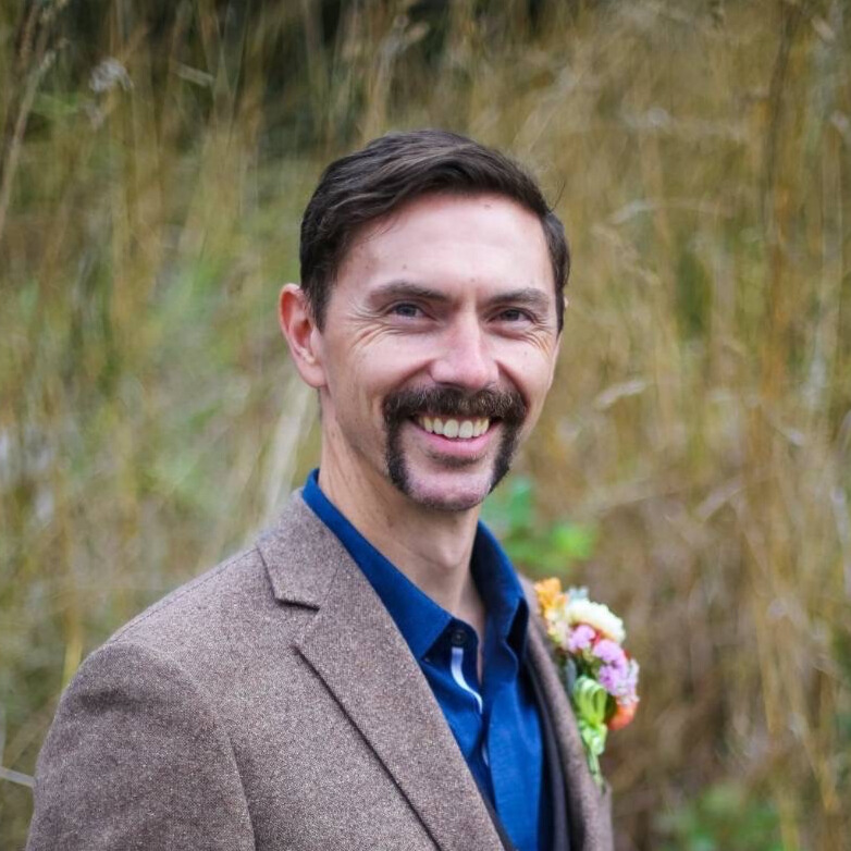 Person with brown hair wearing a blue shirt and brown jacket standing in front of a field