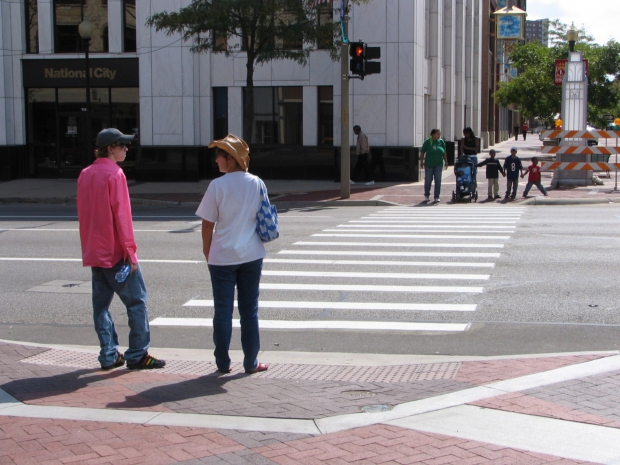 Pedestrians, both adults and children, stand waiting on opposite sides of a crosswalk as the traffic signal glows red.
