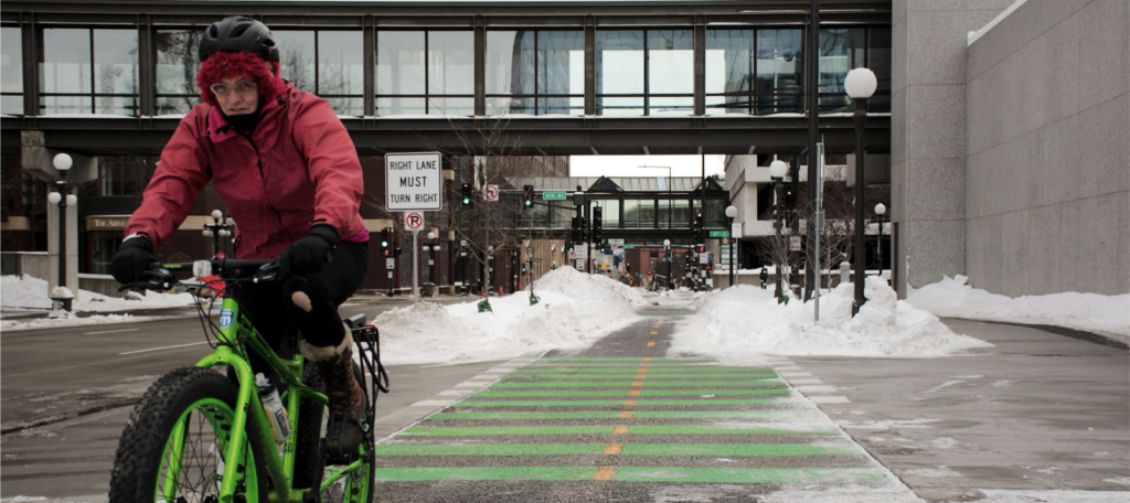 A woman dressed warmly, riding a bike across a shared crosswalk for walking and biking. Behind her the path is partially covered by snow but still usable.