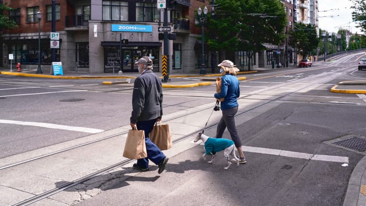 Two people carrying grocery bags with a dog crossing a street in a crosswalk